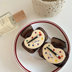 Two art palette hair claw clips are arranged on a white plate with a red rim, with a bottle of perfume in the background.