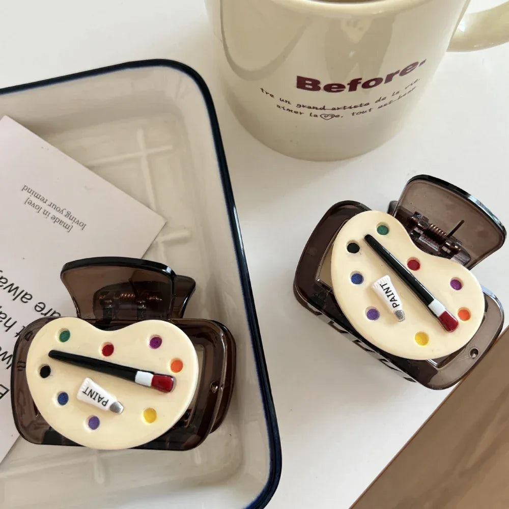 Two art palette hair claw clips are placed on a white tray with some cards, and a mug with the word "Before" is visible in the background.