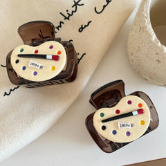 Two art palette hair claw clips are positioned on a beige cloth with handwritten text, alongside a textured white object.