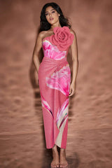 Woman in pink one-piece swimsuit with floral print and large pink flower embellishment on shoulder, standing with hand on hip.