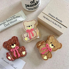 Three teddy bear hair clips in brown, beige, and cream colors, arranged with a tube of hand cream and a box.