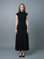 Woman in a black mock neck cap sleeve A-line dress standing against a grey background.