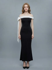 Woman wearing a black off-the-shoulder mermaid gown with a white sash and bow detail, standing against a grey background.