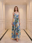 Woman wearing a long, sleeveless maxi dress with a colorful fruit and floral print, standing in a hallway.