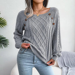 Woman wearing a grey cable knit sweater with buttons, lifting the hem of the sweater, paired with light wash jeans, standing in front of a white wall with a plant.