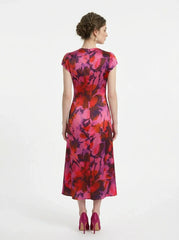 Back view of a woman wearing a pink and red floral print satin midi dress with short sleeves and ruched detailing, standing against a white background.
