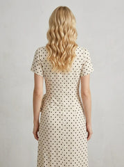 Back view of a woman wearing a cream and black polka dot midi wrap dress with short sleeves, standing against a white textured wall.