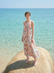 Woman wearing a white floral print maxi dress with a halter neckline and tiered skirt, standing on a rock in the ocean.