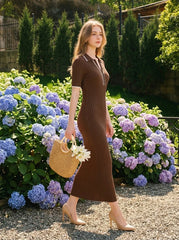 Woman walking in a garden wearing a brown ribbed knit dress with white trim on the collar and sleeves, holding a straw bag with flowers, and wearing nude high heels.