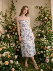 Woman wearing a floral print midi dress with tie shoulder straps and a high slit, standing amongst flowers.