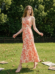 Woman wearing a cream and coral floral print halter midi dress, walking in a grassy field with trees in the background.