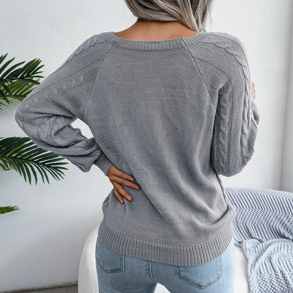 Back view of a woman wearing a grey cable knit sweater with buttons, paired with light wash jeans, standing in front of a white wall with a plant and a couch.