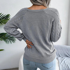 Back view of a woman wearing a grey cable knit sweater with buttons, paired with light wash jeans, standing in front of a white wall with a plant and a couch.