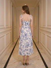 Back view of a woman wearing a floral print midi dress with tie shoulder straps and a high slit, standing in a hallway.
