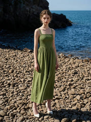 Woman wearing a green linen midi dress with a smocked bodice and square neckline, posing on a pebble beach with the ocean in the background.
