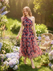 Woman wearing a navy floral print one-shoulder dress with a tiered skirt and ruffle detail, standing in a garden with hydrangeas and flowers.