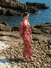 woman wearing a pink and red floral print satin maxi wrap dress with long bishop sleeves and a v-neck, posing on a rocky beach with the ocean in the background