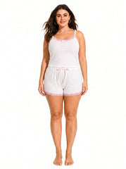 Woman wearing a white ribbed camisole and shorts pajama set with pink lace trim and bow accents.