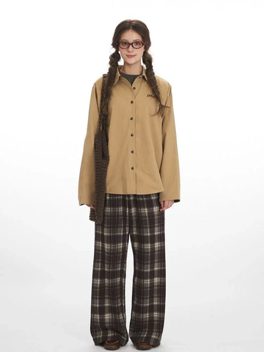 Model wearing a tan button-down shirt and brown plaid wide-leg pants, holding a brown knitted bag.
