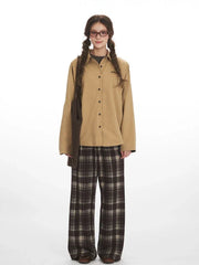Model wearing a tan button-down shirt and brown plaid wide-leg pants, holding a brown knitted bag.