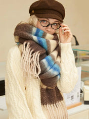 Close-up of a model wearing a cream cable knit sweater and a brown plaid scarf with cream, blue, and brown stripes. She is also wearing a brown flat cap and round glasses.