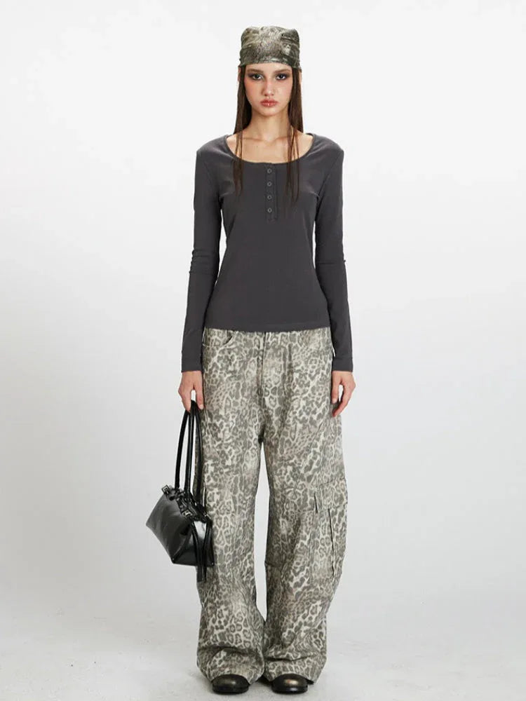 Woman wearing a dark grey long-sleeve top with a square neckline and button details, paired with wide-leg grey leopard print pants and a dark handbag. She is also wearing a patterned headscarf.