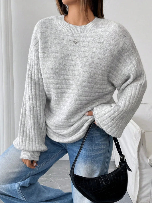 Woman wearing a light grey oversized knit sweater with balloon sleeves and a crew neck, paired with blue denim jeans and a black handbag.