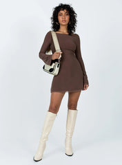 Woman wearing a long sleeve brown mini dress, white crossbody bag, and white boots against a white background.