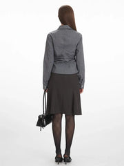 Back view of a woman wearing a grey long-sleeved button-down shirt with ruching detail, a black skirt, black tights, and black heels, holding a black handbag.