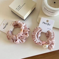 Two light pink satin scrunchies with ruffled texture are displayed on a white surface. One scrunchie has a wooden bead detail, and the other has a pearl embellishment.