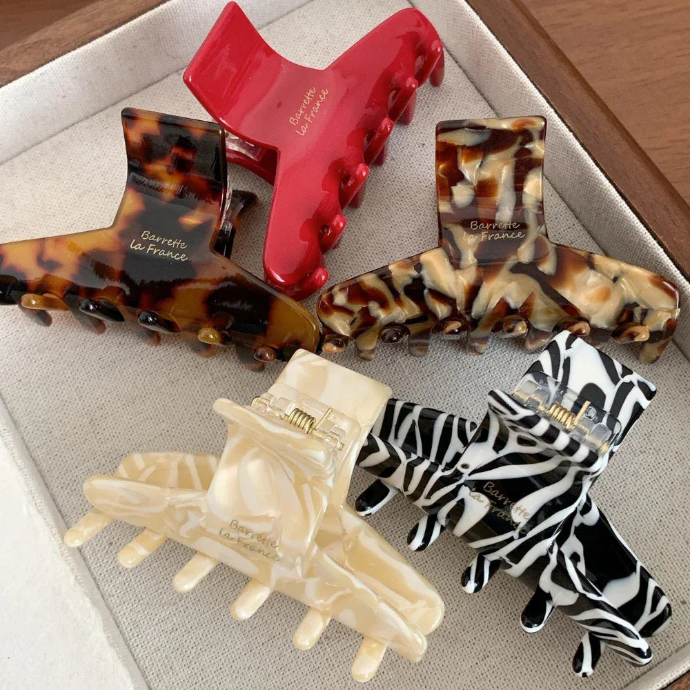 A top-down view of five hair claw clips on a textured white surface. Patterns include tortoiseshell, marbled cream, zebra print, and solid red.
