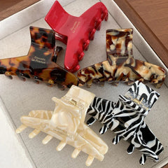 A top-down view of five hair claw clips on a textured white surface. Patterns include tortoiseshell, marbled cream, zebra print, and solid red.