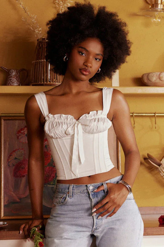 Woman wearing a white ruched corset top with a bow accent and light wash denim jeans, standing with hands in pockets.