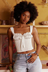 Woman wearing a white ruched corset top with a bow accent and light wash denim jeans, standing with hands in pockets.