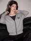 Woman wearing a grey knit cardigan with black trim and embroidery, layered over a dark top, paired with dark wide-leg jeans, sitting on a black couch.