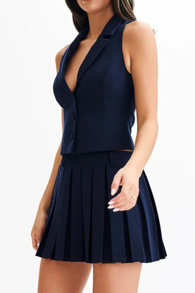 Woman in a navy blue sleeveless blazer vest and pleated skirt, with hand on hip.