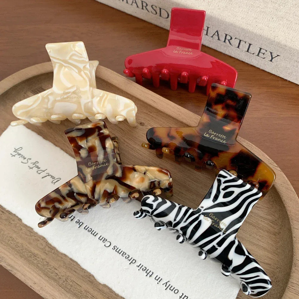 A flat lay of five hair claw clips on a wooden tray. Patterns include tortoiseshell, marbled cream, zebra print, and solid red.