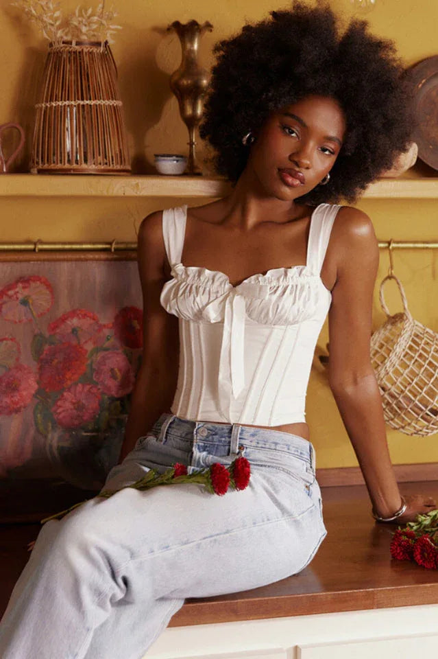 Woman wearing a white ruched corset top with a bow accent and light wash denim jeans, sitting on a counter.