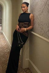 Woman in a floor-length black sleeveless dress with sheer lace waist detailing and a high neckline, holding a handbag.