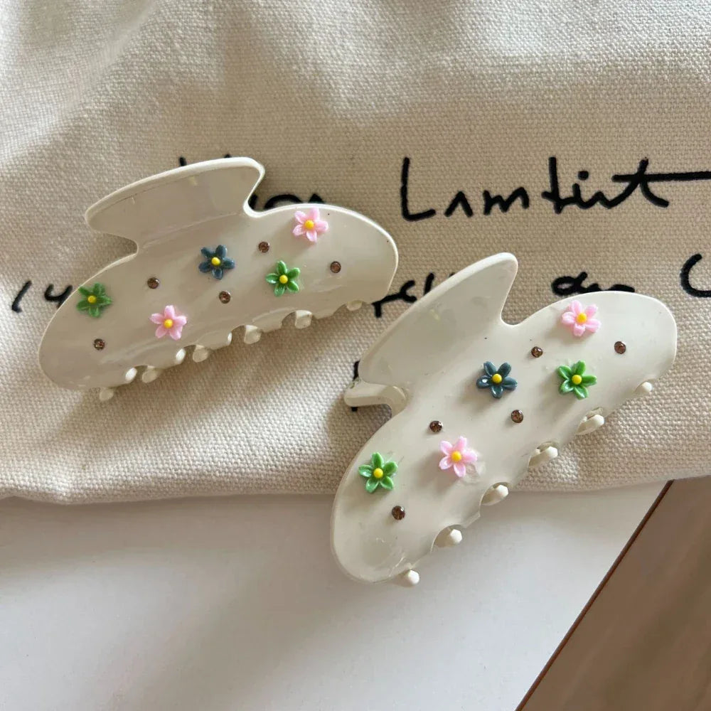 Two cream-colored claw clips with floral and gemstone embellishments, resting on a textured fabric.