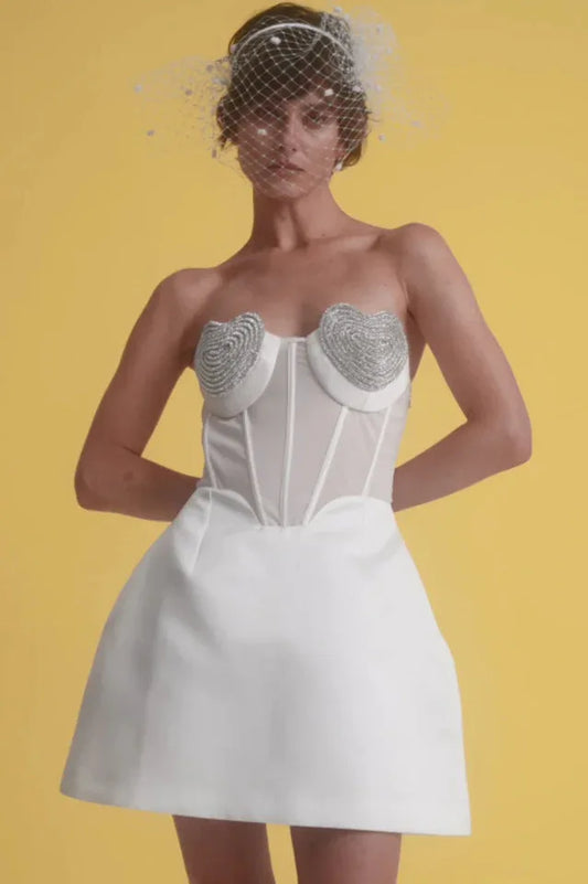 Model wearing a white strapless dress with heart-shaped embellishments on the bust, a corset bodice, and a full skirt, against a yellow background.