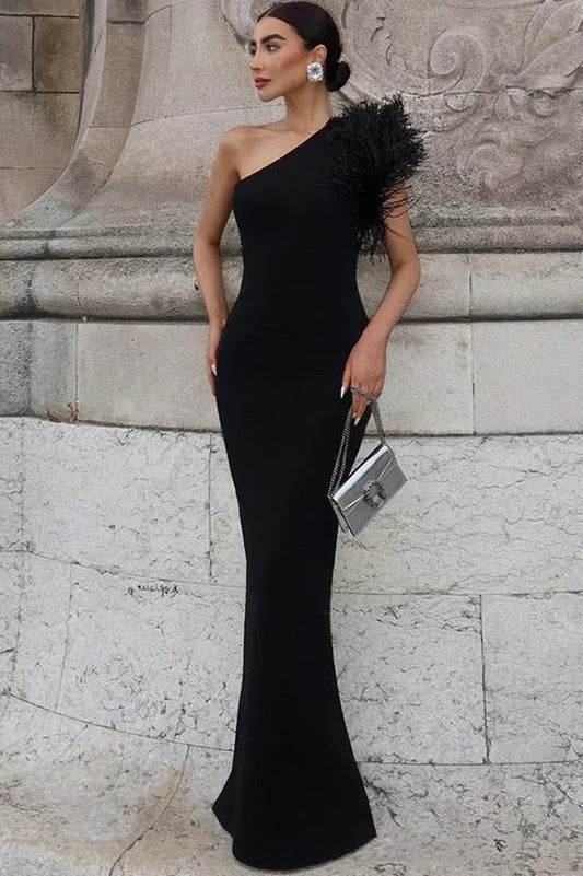 Woman wearing a black one-shoulder crepe gown with a feather accent on the shoulder and a mermaid hemline, standing in front of a stone wall.