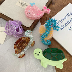 A flat lay of acrylic hair clips featuring a pink whale, a purple octopus, a blue seahorse, and two turtles arranged on a wooden surface and white paper.