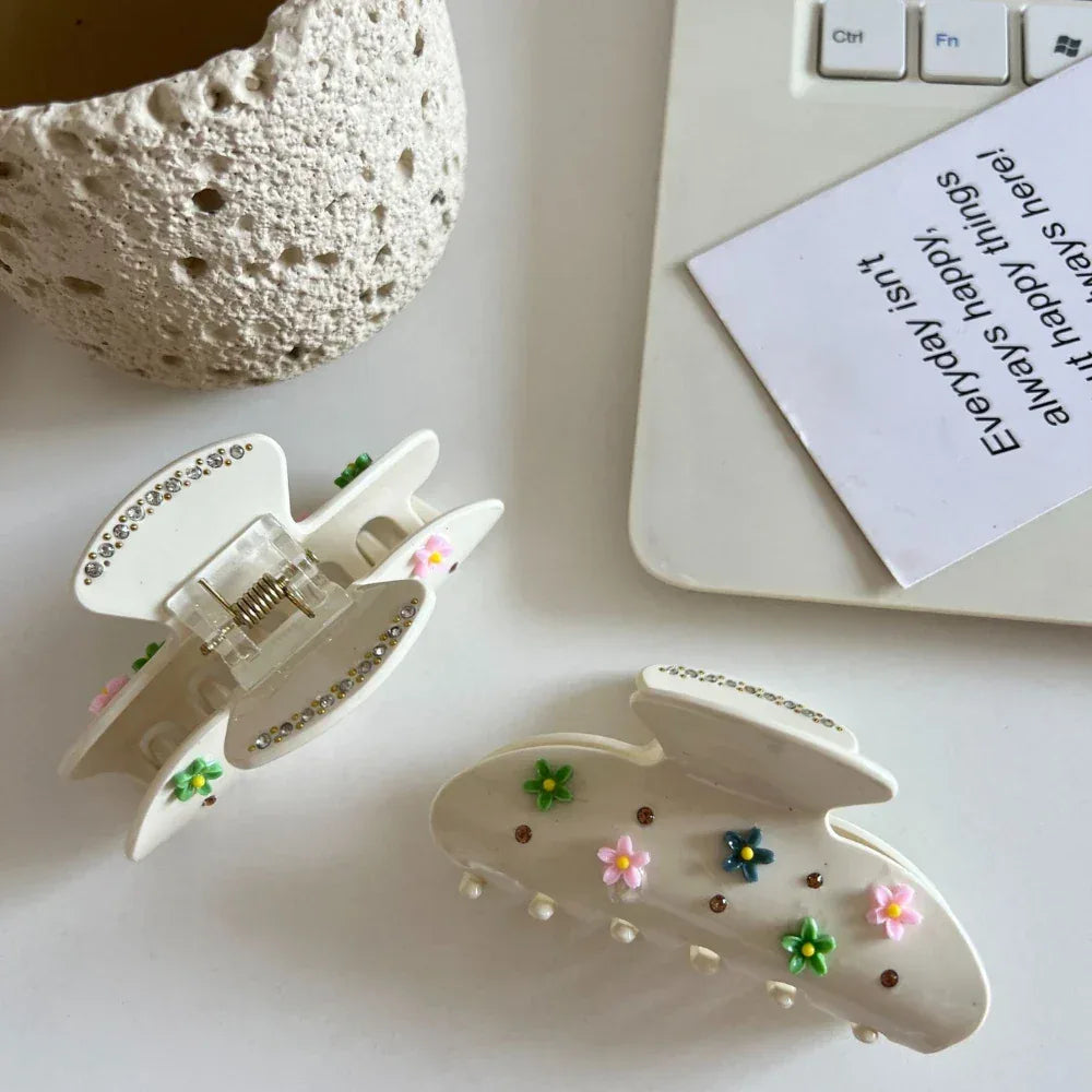 Two cream-colored claw clips with floral and gemstone embellishments, placed on a white surface next to a laptop.