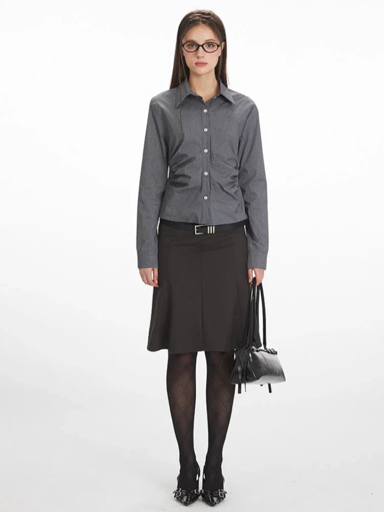 Woman wearing a grey long-sleeved button-down shirt with ruching detail, a black skirt, black tights, and black heels, holding a black handbag.