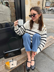 Woman sitting on a bench outside a cafe, holding a coffee cup and wearing a striped knit sweater and blue jeans.