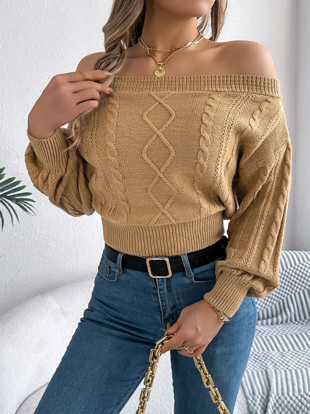 Back view of a woman wearing a tan off-the-shoulder cable knit cropped sweater and blue jeans.