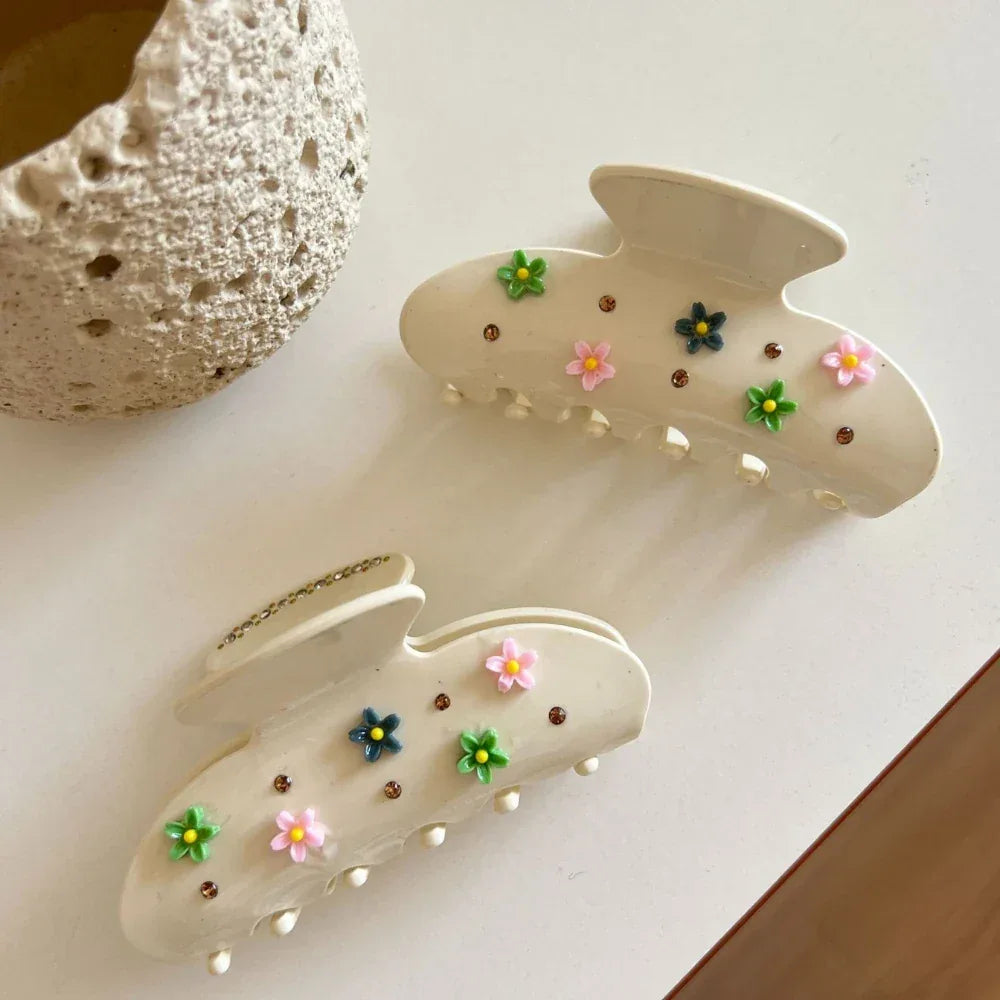 Two cream-colored claw clips with floral and gemstone embellishments, viewed from a top-down angle on a white surface.