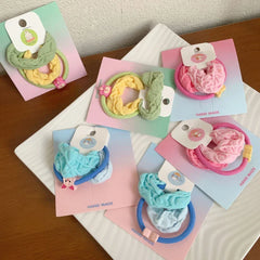 A variety of handmade scrunchie sets in soft pastel shades, each packaged on a card with a different cute charm.