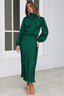 Woman wearing a long-sleeved emerald green satin dress with a cowl neckline and a ruffled hem, standing in a studio.
